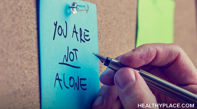 Hand holding a pen writing "you are not alone" on a blue sticky note.